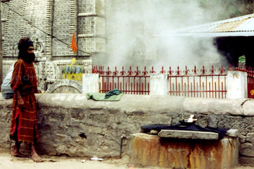Ein indischer Pilger vor einem Tempel im Himalaya mit heißer Quelle. Im Hintergrund rechts im Bild erkennt man den aufsteigenden Wasserdampf.
